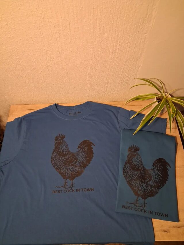 A blue "Best Cock in Town" rooster shirt on a table, a second one folded beside it