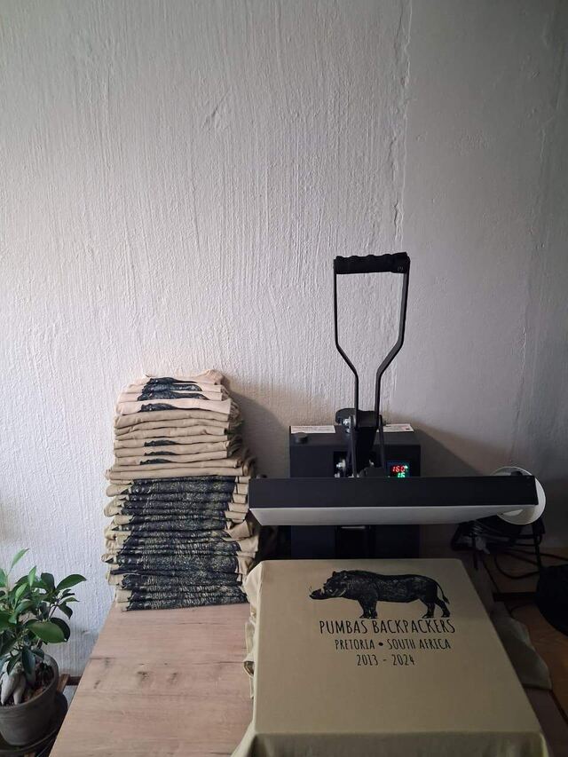 A heat press beside a stack of printed Pumbas Backpackers shirts